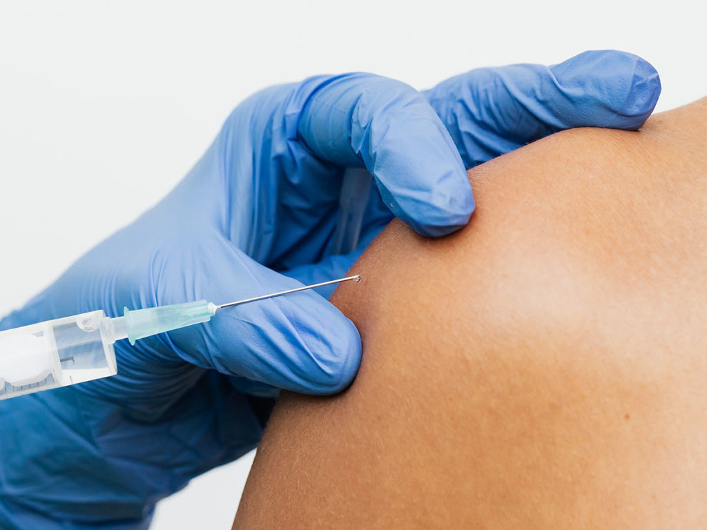 Die Nadel einer Impf-Spritze vor der Schulter einer Patientin