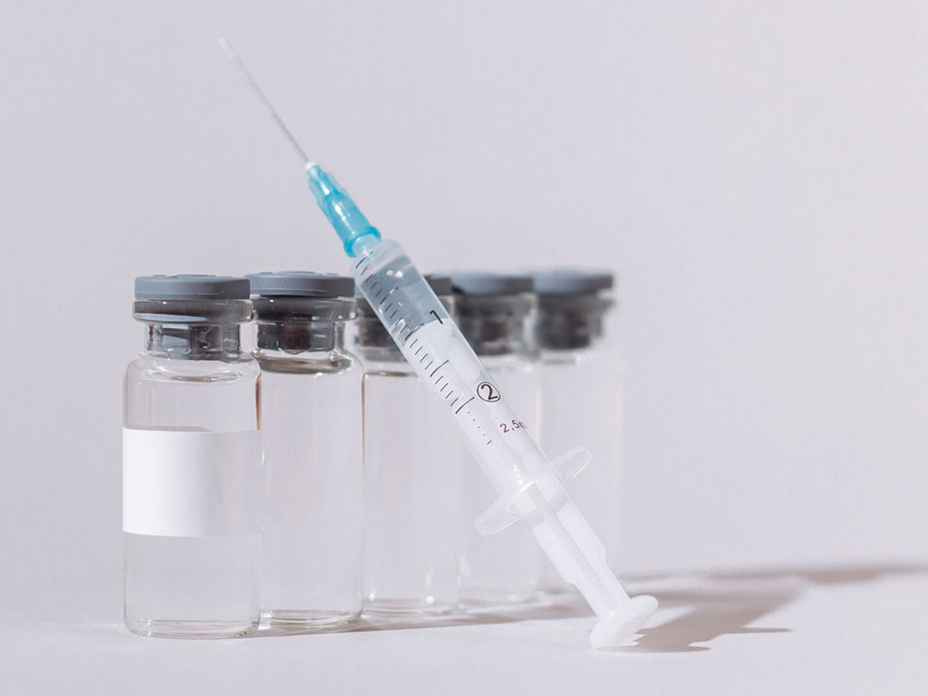 Eine Impf-Spritze vor einigen Impfstoff-Fläsch­chen